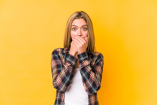 Giovane donna bionda caucasica isolata su sfondo giallo scioccato che copre la bocca con le mani.