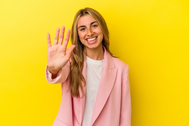 Giovane donna bionda caucasica isolata su sfondo giallo sorridente allegro che mostra il numero cinque con le dita.