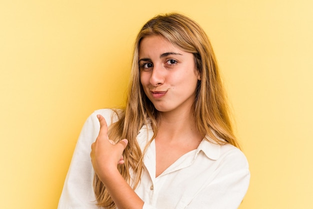 Giovane donna bionda caucasica isolata su sfondo giallo sorridente e puntata da parte, mostrando qualcosa in uno spazio vuoto.