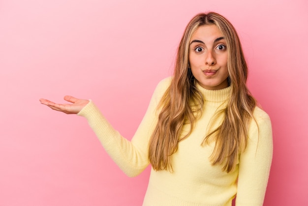 Giovane donna bionda caucasica isolata su sfondo rosa che mostra uno spazio di copia su un palmo e tenendo un'altra mano sulla vita.