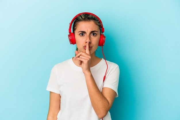 Giovane donna bionda che ascolta la musica sulle cuffie isolate sulla parete blu mantenendo un segreto o chiedendo il silenzio