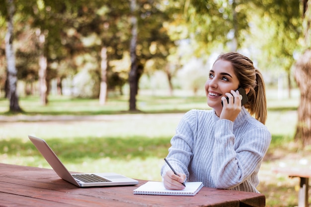 Giovane donna bionda che comunica sul telefono mentre prendendo le note all&#39;esterno.