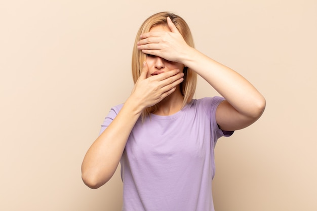 Giovane donna bionda che copre il viso con entrambe le mani dicendo no alla telecamera!