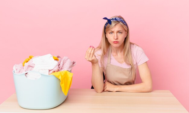 Giovane donna bionda che fa un gesto capice o denaro, dicendoti di pagare. lavare i vestiti concetto clothes