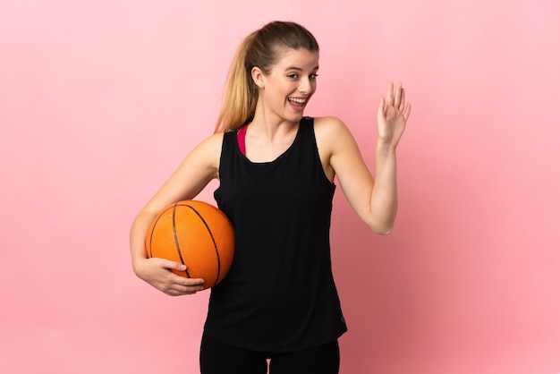 Giovane donna bionda che gioca a basket isolato su spazio rosa salutando con la mano con felice espressione