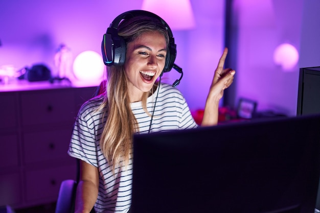 Giovane donna bionda che gioca ai videogiochi sorridendo felice indicando con la mano e il dito di lato