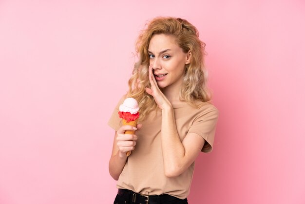 Giovane donna bionda che giudica un gelato della cornetta isolato sulla parete rosa che bisbiglia qualcosa