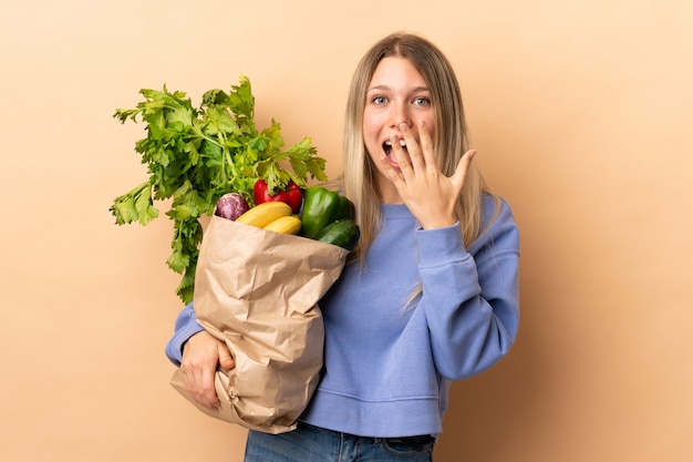 Giovane donna bionda che giudica una borsa piena delle verdure sopra la parete isolata con espressione facciale di sorpresa