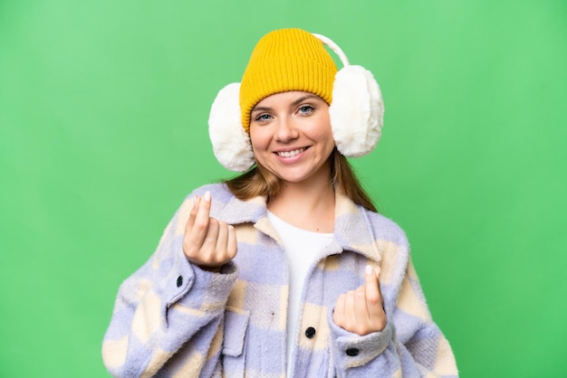 Giovane donna bionda che indossa manicotti invernali su sfondo chroma key isolato che fa gesto di denaro