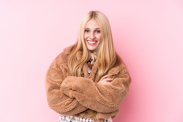 Giovane donna bionda che indossa un cappotto contro un muro rosa che si sente fiducioso, incrociando le braccia con determinazione.