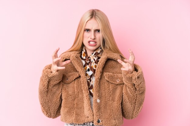 Giovane donna bionda che indossa un cappotto contro un muro rosa sconvolto urlando con le mani tese.