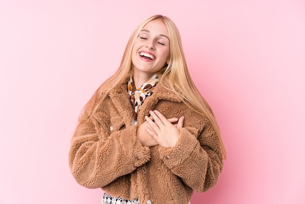 Giovane donna bionda che indossa un cappotto contro una parete rosa che ride tenendo le mani sul cuore