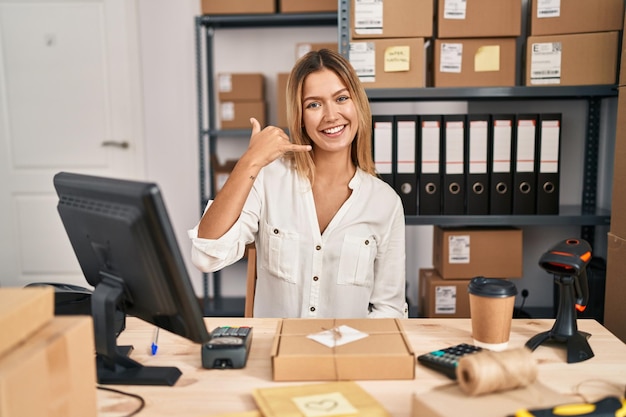 Giovane donna bionda che lavora presso un e-commerce di piccole imprese che sorride facendo il gesto del telefono con la mano e le dita come parlare al telefono. comunicare concetti.