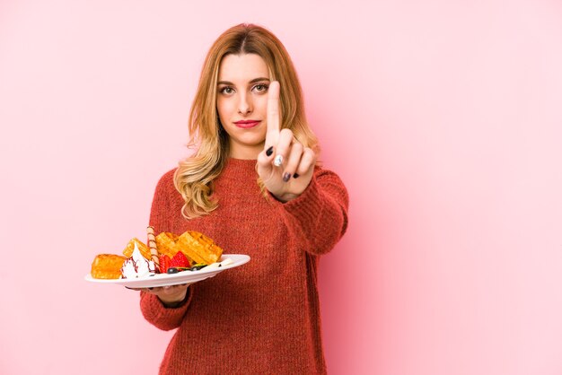 Giovane donna bionda che mangia un dessert della cialda isolato che mostra il numero uno con il dito.