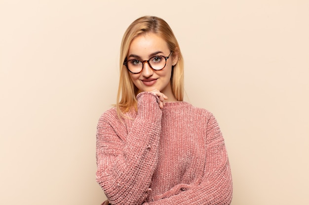 Giovane donna bionda che sembra felice e sorridente con la mano sul mento, chiedendosi o facendo una domanda, confrontando le opzioni