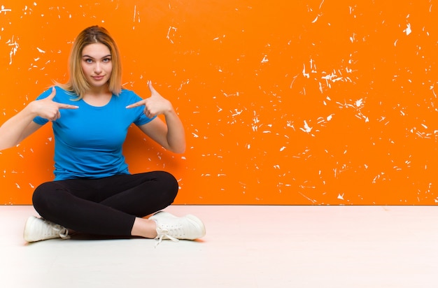 Giovane donna bionda che sembra fiero, positivo e casuale che indica il petto con entrambe le mani che si siedono sul pavimento