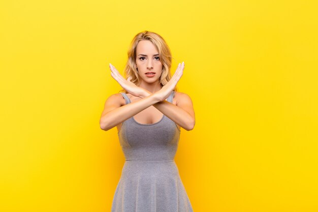 Giovane donna bionda che sembra infastidita e stanca del tuo atteggiamento, dicendo abbastanza! le mani incrociate sul davanti, dicendoti di fermarti contro il muro arancione