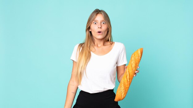 Giovane donna bionda che sembra molto scioccata o sorpresa e tiene in mano una baguette di pane