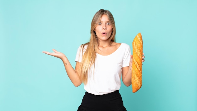 Giovane donna bionda che sembra sorpresa e scioccata, con la mascella caduta in possesso di un oggetto e con in mano una baguette di pane