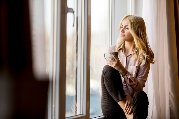 Giovane donna bionda che si rilassa e che beve caffè dalla finestra