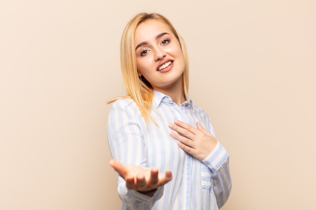 Giovane donna bionda che si sente felice e innamorata, sorridendo con una mano vicino al cuore e l'altra allungata in avanti