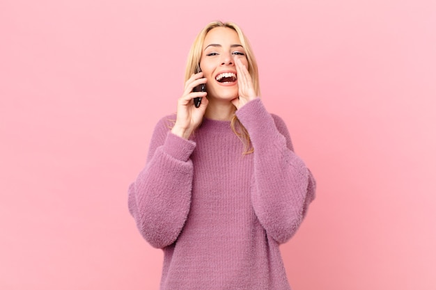 Giovane donna bionda che si sente felice, fa un grande grido con le mani vicino alla bocca e parla con uno smartphone