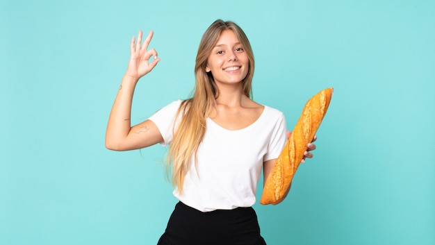 Giovane donna bionda che si sente felice, mostra approvazione con un gesto ok e tiene in mano una baguette di pane