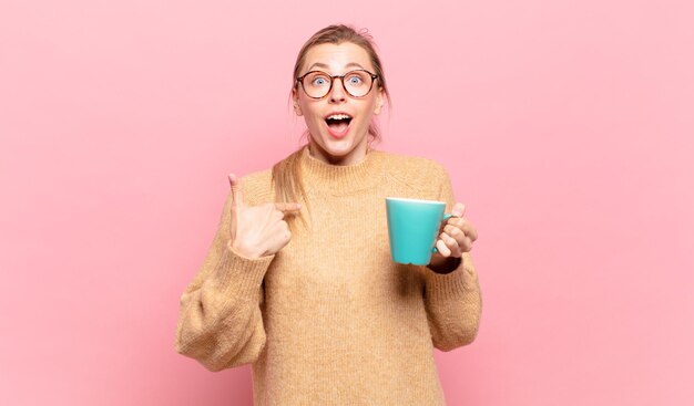 Giovane donna bionda che si sente felice, sorpresa e orgogliosa, indicando se stessa con uno sguardo eccitato e stupito. concetto di caffè