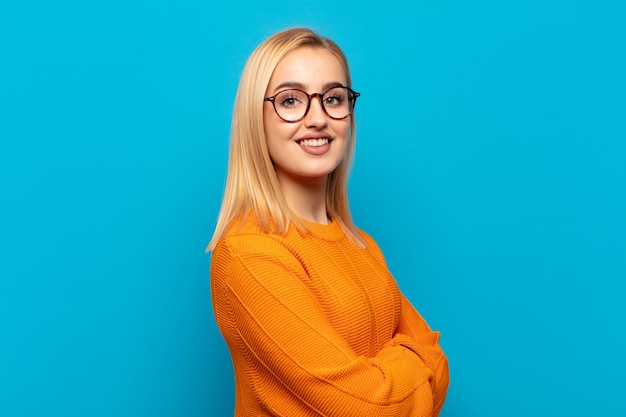 Giovane donna bionda che sorride alla macchina fotografica con le braccia incrociate e un'espressione felice, fiduciosa, soddisfatta, vista laterale