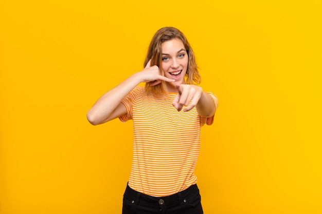 Giovane donna bionda che sorride allegramente e indicando mentre ti fa una chiamata in seguito gesto, parlando al telefono contro la parete di colore piatto