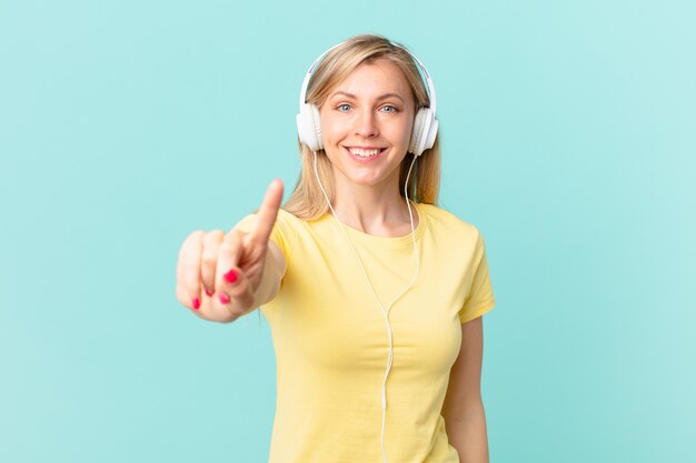 Giovane donna bionda che sorride con orgoglio e fiducia facendo il numero uno e ascoltando musica.