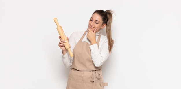 Giovane donna bionda che sorride con un'espressione felice e sicura con la mano sul mento, chiedendosi e guardando di lato