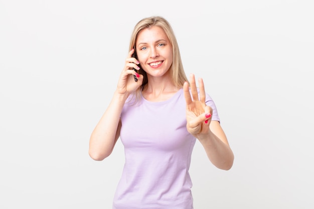 Giovane donna bionda che sorride e sembra amichevole, mostra il numero tre e chiama con uno smartphone
