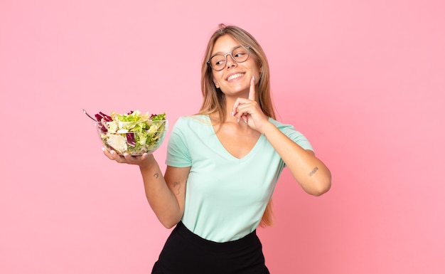 Giovane donna bionda che sorride felice e sogna ad occhi aperti o dubita e tiene in mano un'insalata