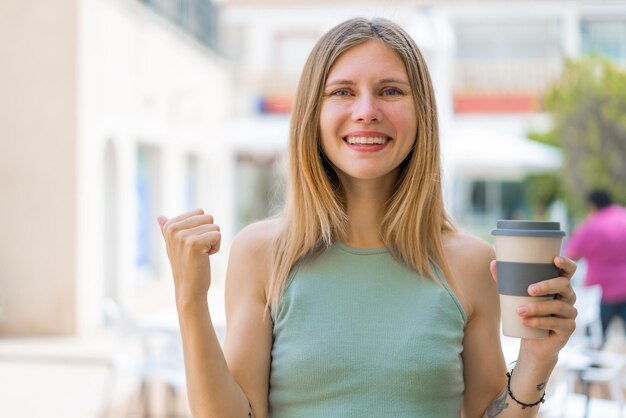 Giovane donna bionda che tiene un caffè da asporto all'aperto che punta verso il lato per presentare un prodotto
