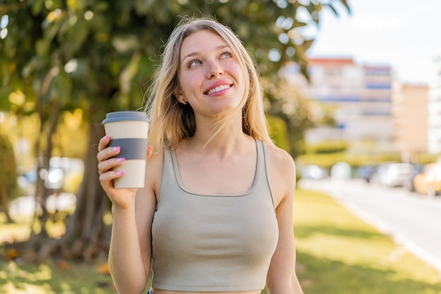 Giovane donna bionda che tiene un caffè da asporto all'aperto guardando in alto mentre sorride