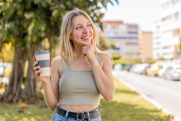 Giovane donna bionda che tiene un caffè da asporto all'aperto pensando un'idea e guardando un lato