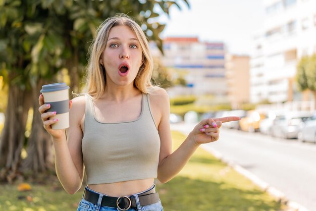 Giovane donna bionda che tiene un caffè da asporto all'aperto sorpreso e che indica il lato