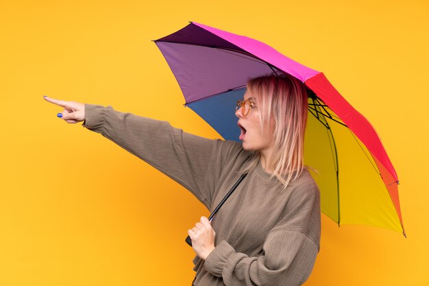 Giovane donna bionda che tiene un ombrello sopra la parete gialla isolata che indica dito il lato