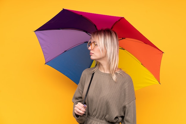 Giovane donna bionda che tiene un ombrello sopra la parete gialla isolata che sembra laterale