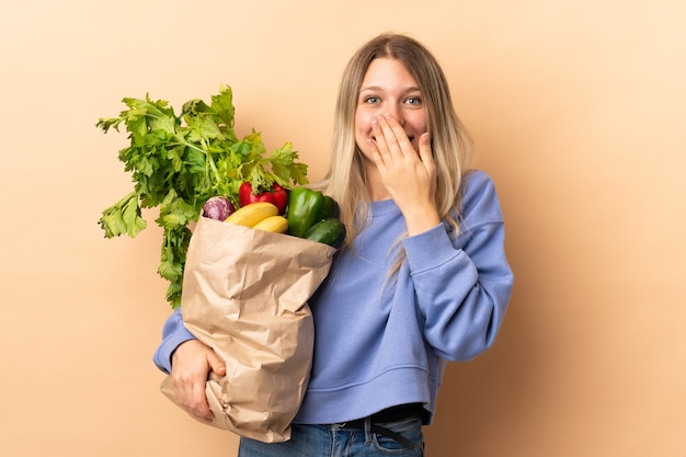 Giovane donna bionda che tiene un sacchetto pieno di verdure sopra la parete isolata con l'espressione facciale di sorpresa