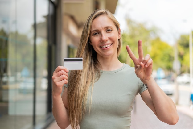 Giovane donna bionda che tiene una carta di credito all'aperto sorridendo e mostrando il segno della vittoria