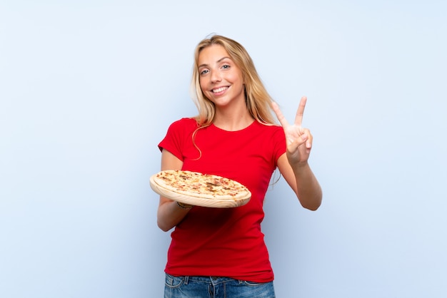 Giovane donna bionda che tiene una pizza sopra la parete blu isolata che sorride e che mostra il segno di vittoria