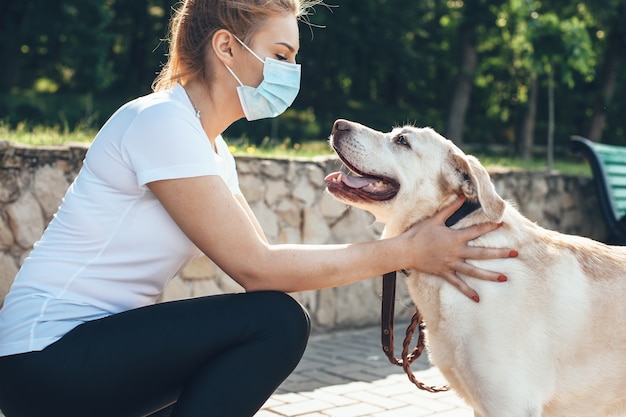 Giovane donna bionda che tocca il suo cane mentre indossa la mascherina medica sul viso e cammina nel parco