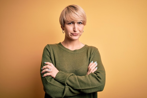 Giovane donna bionda con capelli corti moderni che indossa un maglione casuale su sfondo giallo scettica e nervosamente disapprovante espressione sul viso con le braccia incrociate persona negativa