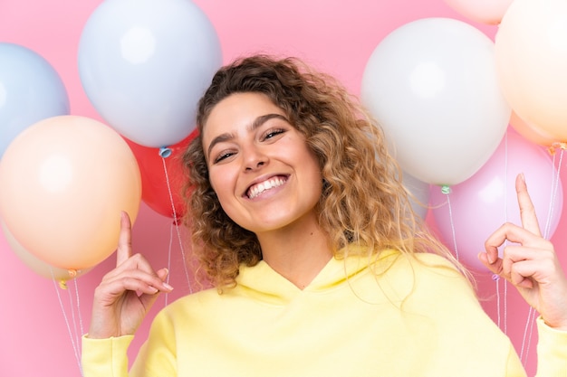 Giovane donna bionda con capelli ricci che cattura molti palloncini isolati in rosa