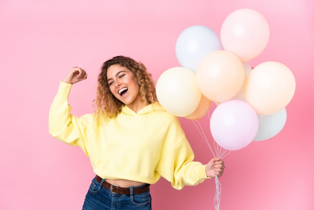 Giovane donna bionda con capelli ricci che cattura molti palloni sulla parete rosa che celebra una vittoria