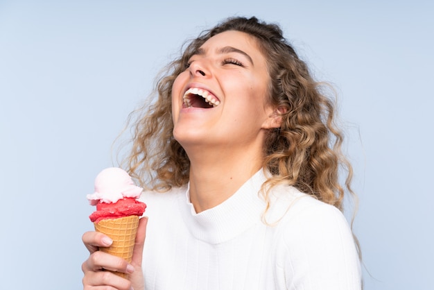 Giovane donna bionda con capelli ricci che giudicano un gelato della cornetta isolato sulla parete blu