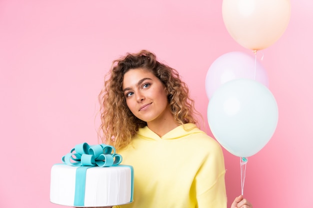 Giovane donna bionda con capelli ricci che prende molti palloni e che tiene una grande torta sul rosa