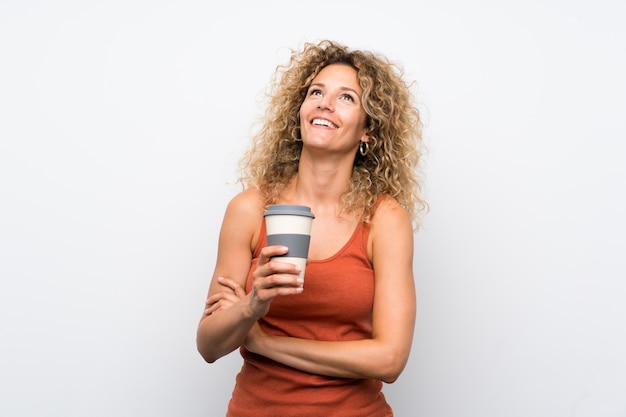 Giovane donna bionda con capelli ricci che tengono un caffè da asporto che osserva in su mentre sorridendo